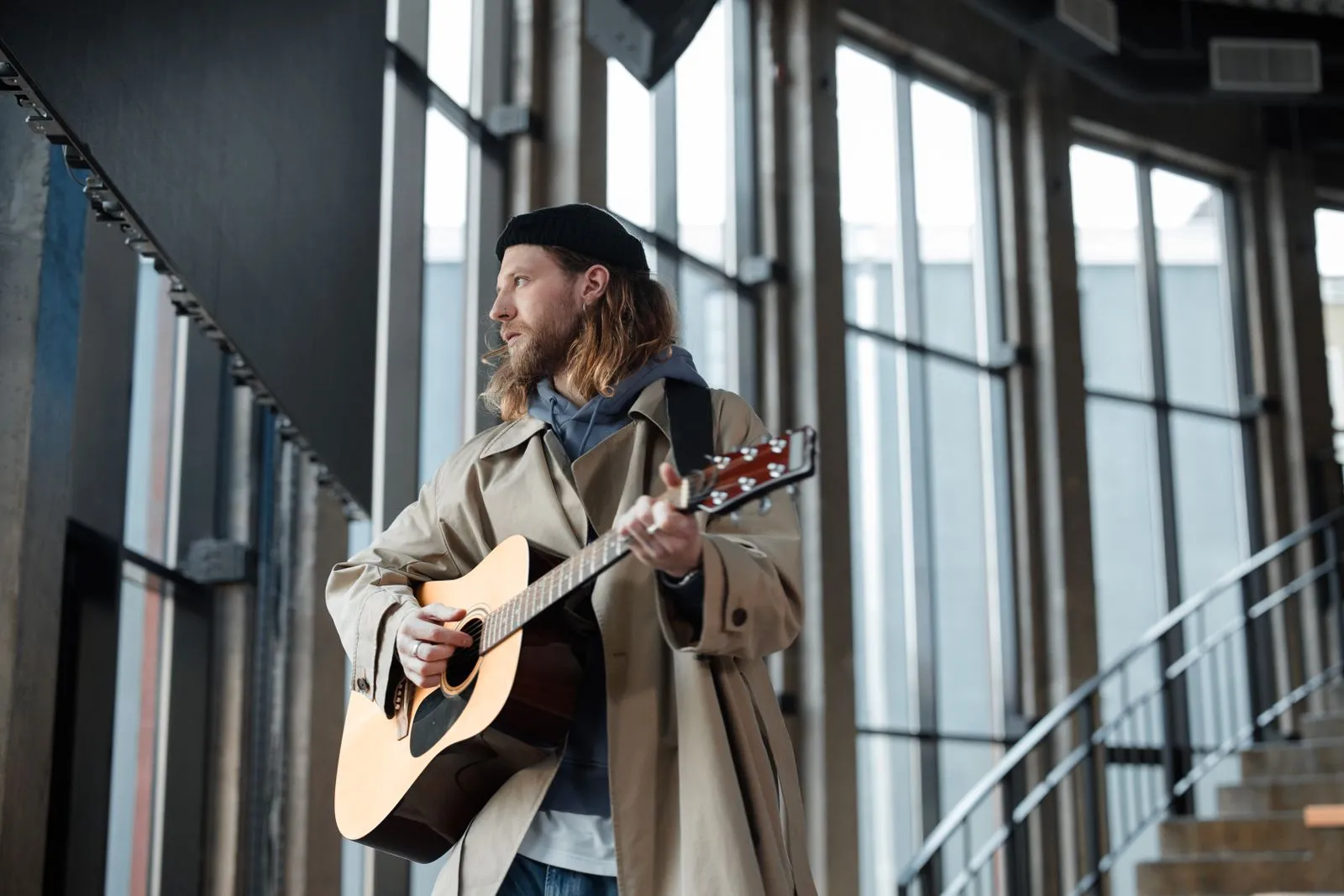 Male musician playing an acoustic guitar indoors near large windows.